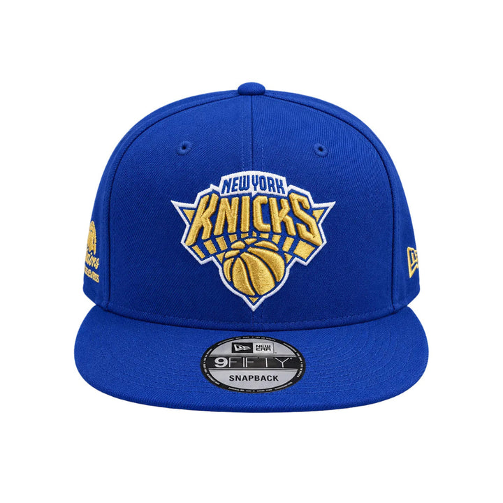 A royal blue New York Knicks snapback cap featuring a gold and white embroidered team logo on the front, with a visible New Era 9FIFTY sticker on the brim.